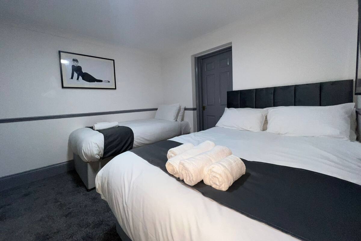 A hotel room with two single beds, a large bed, and a gray headboard. There is a framed picture on the wall and a door to the right.