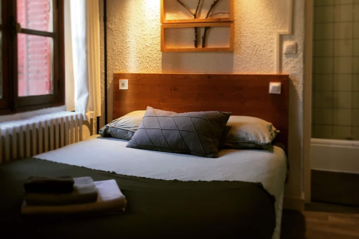 Una cama bien hecha con un cabecero de madera, una mesita de noche con una lámpara y una ventana con cortinas.