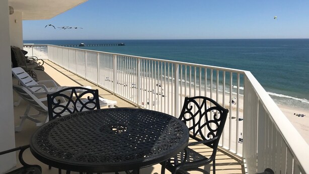 A balcony with a table and chairs overlooking a beach with a pier in the distance. - Lighthouse 801 Corner Unit Beachfront w HUGE wrap balcony! Book for Aug., Fall! (Gulf Shores)