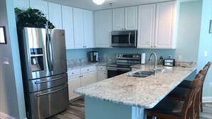 A modern kitchen with white cabinets, stainless steel appliances, a granite countertop, and a central island with bar stools. - Lighthouse 801 Corner Unit Beachfront w HUGE wrap balcony! Book for Aug., Fall! (Gulf Shores)