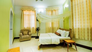 A bedroom with a bed, a chair, a bedside table, a standing fan, and a ceiling fan. - Mama Thea Homes - Hostel (Moshi)
