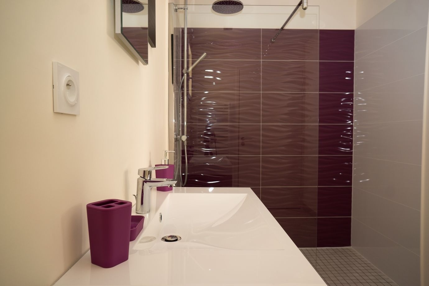 Un baño moderno con un lavamanos blanco, un dispensador de jabón morado y una cabina de ducha de cristal.