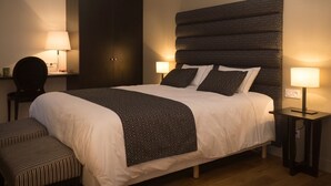 Ein ordentlich bezogenes Bett mit einem gemusterten Kopfteil, zwei Nachttischlampen, einem dunklen Kleiderschrank und einer gestreiften Bank.