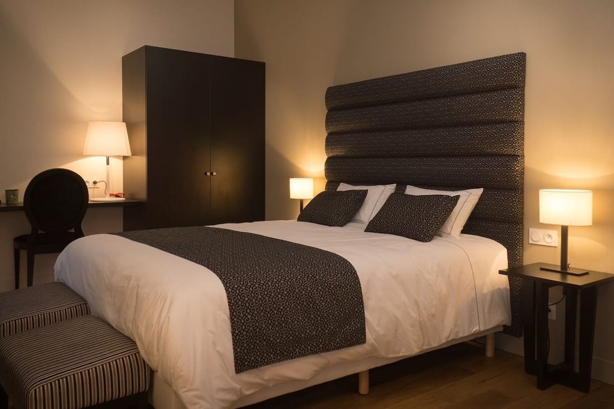 A neatly made bed with a patterned headboard, two bedside lamps, a dark wardrobe, and a striped bench.