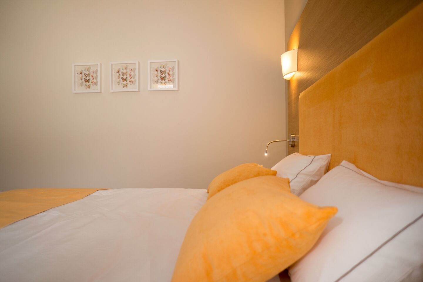 Una cama bien hecha con almohadas blancas y naranjas, un cabecero de madera y tres cuadros en la pared.