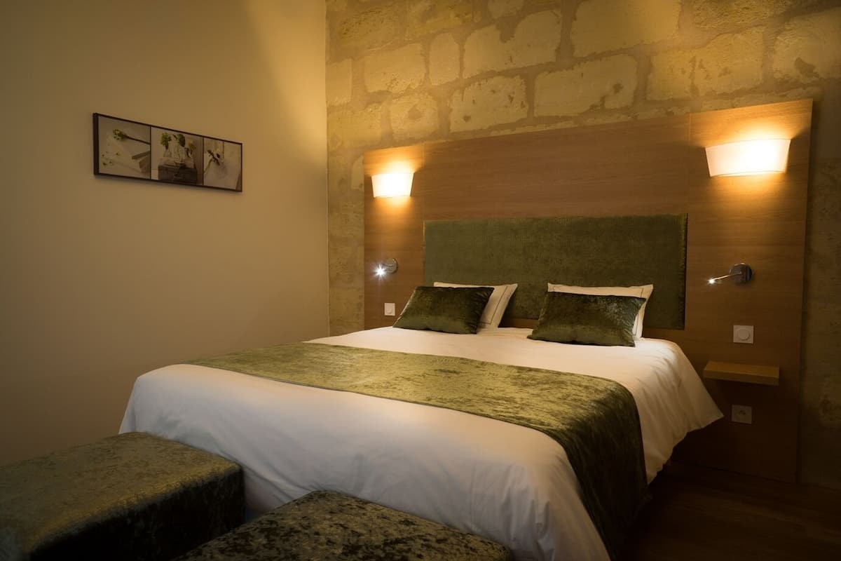A neatly made bed with a green blanket and pillows, a wooden headboard, and two wall-mounted lights.