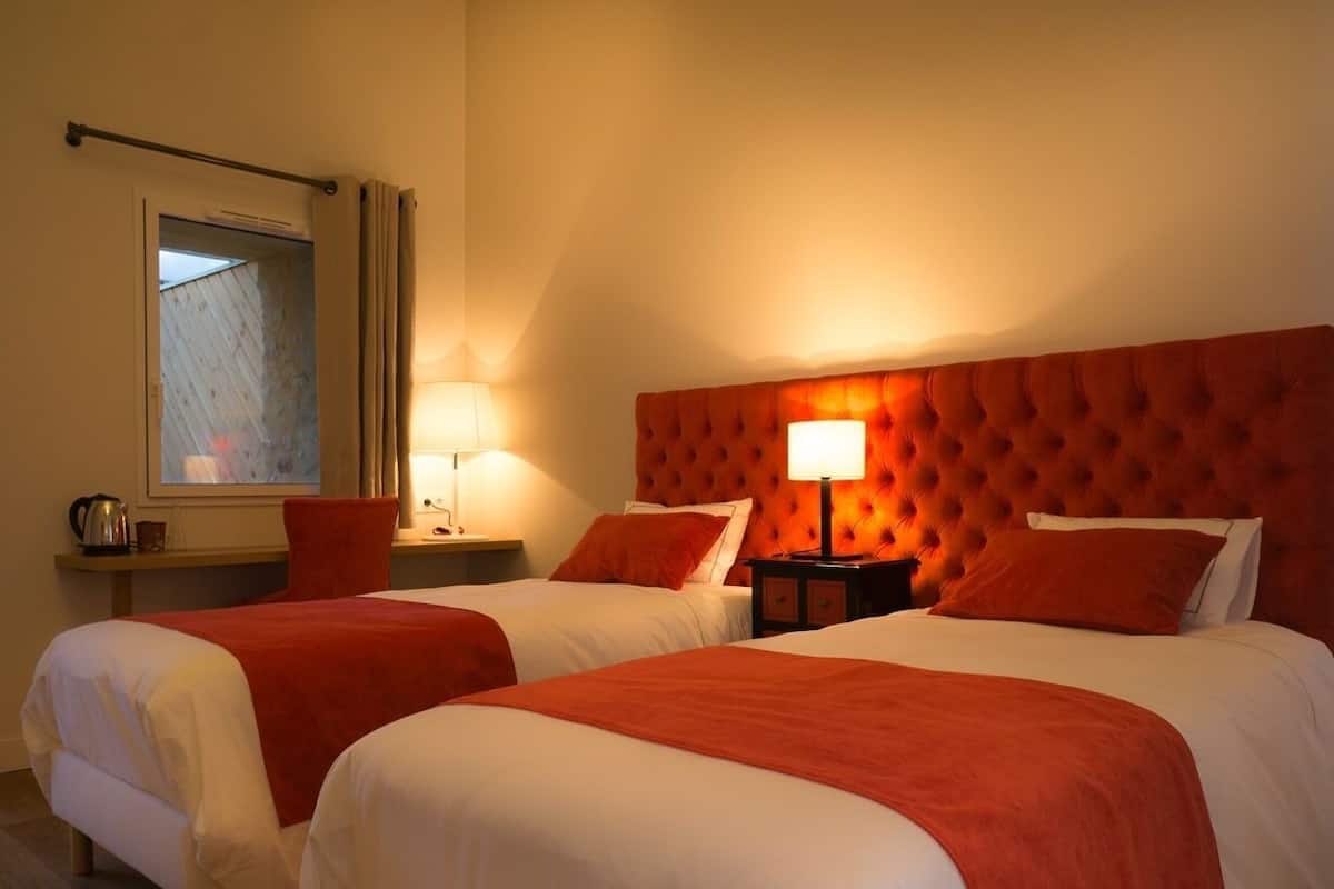 A hotel room with two beds, each with white linens and red accents, a large tufted headboard, and a bedside table with a lamp.