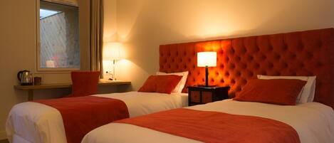 A hotel room with two beds, each with white linens and red accents, a large tufted headboard, and a bedside table with a lamp.