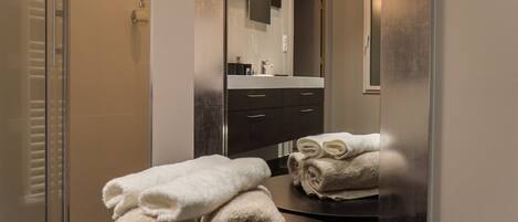A modern bathroom with a glass shower enclosure, a dark vanity with a mirror, and neatly folded towels on a round table.