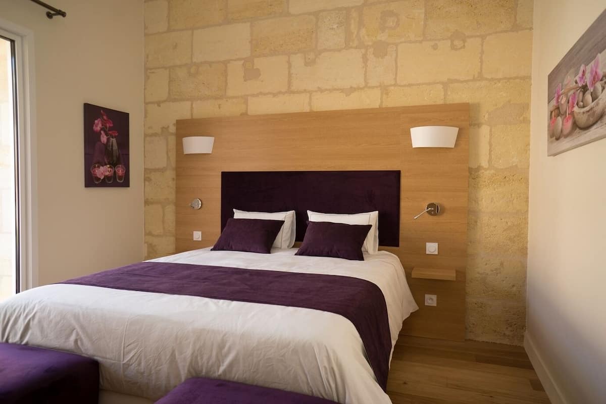 A bedroom with a wooden headboard, two bedside tables, a bed with white and purple bedding, and a brick wall.