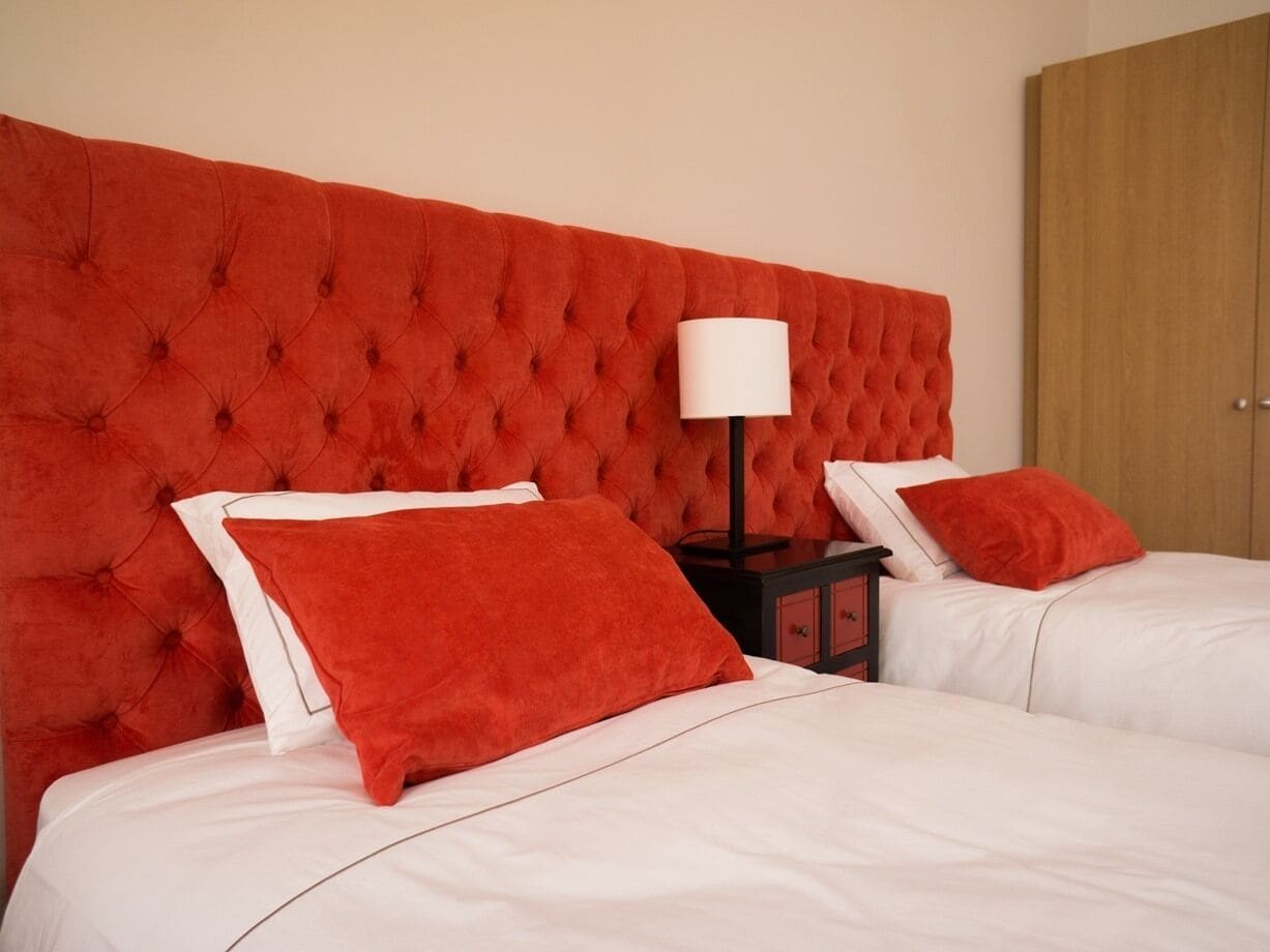 A hotel room with two beds, each with white bedding and red pillows. There is a red tufted headboard, a bedside table with a lamp, and a wooden wardrobe.