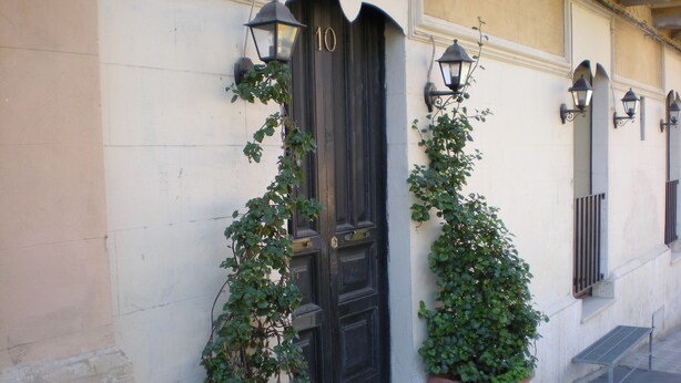 A building facade with a black door marked with the number 10, flanked by two potted plants and two wall-mounted lanterns.