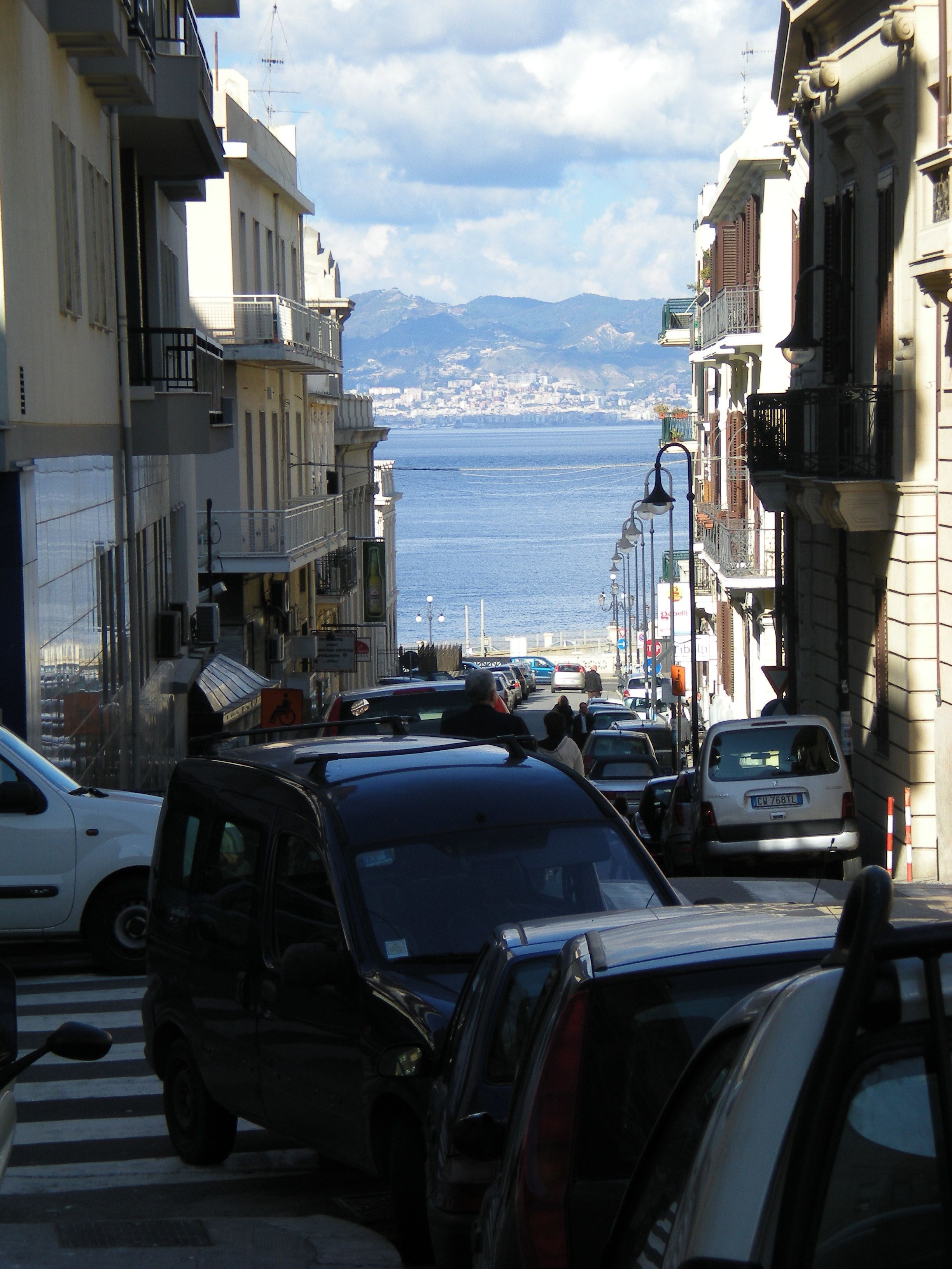 A narrow street with parked cars and buildings on both sides, leading to a body of water with mountains in the distance.