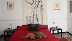 A bedroom with a large bed, a red bedspread, and a large painting of a classical figure on the wall. There are two bedside tables with lamps and framed pictures on the walls.