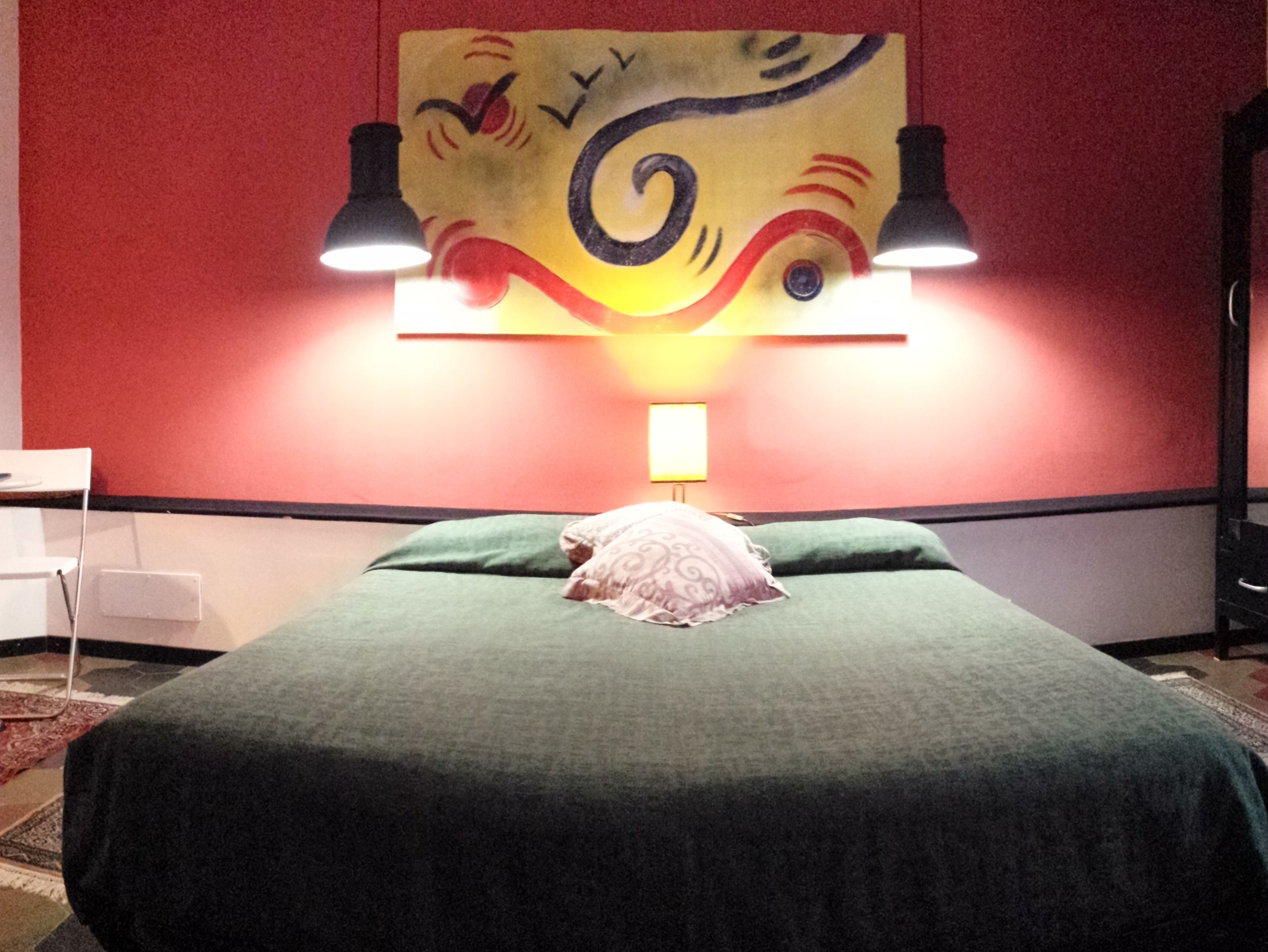 A hotel room with a bed, green pillows, a wall painting, and two wall-mounted lamps.