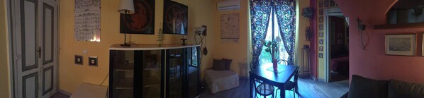 A compact room with a bed, a small table, and a chair. There is a window with a floral curtain, and the walls are decorated with framed pictures.