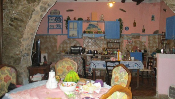 A dining area with a stone archway, a table set with food, chairs, and a kitchen in the background. - L'Antica Locanda (Iglesias)
