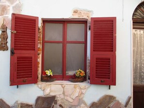 A stone building with a window featuring red shutters and two flower arrangements on the sill. - L'Antica Locanda (Iglesias)