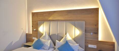 Una habitación de hotel moderna con cabecera de madera, dos camas con ropa blanca, almohadas azules, una mesita de noche con teléfono y un estante pequeño.