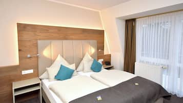 Habitación de hotel con cabecera de madera, cama con ropa blanca y almohadas azules, mesita de noche y ventana con cortinas de gasa.
