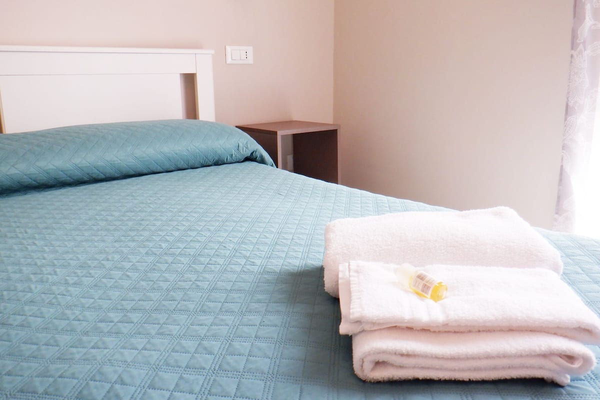 A neatly made bed with a blue quilt, a white pillow, and a stack of white towels with a bottle on top.