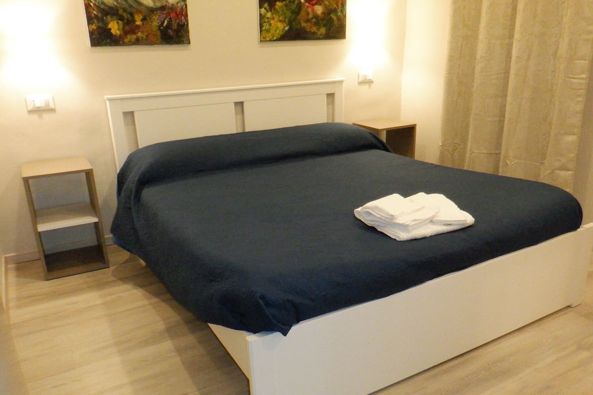 A neatly made bed with a dark blue bedspread and white pillows, a small bedside table, and a wall-mounted shelf.