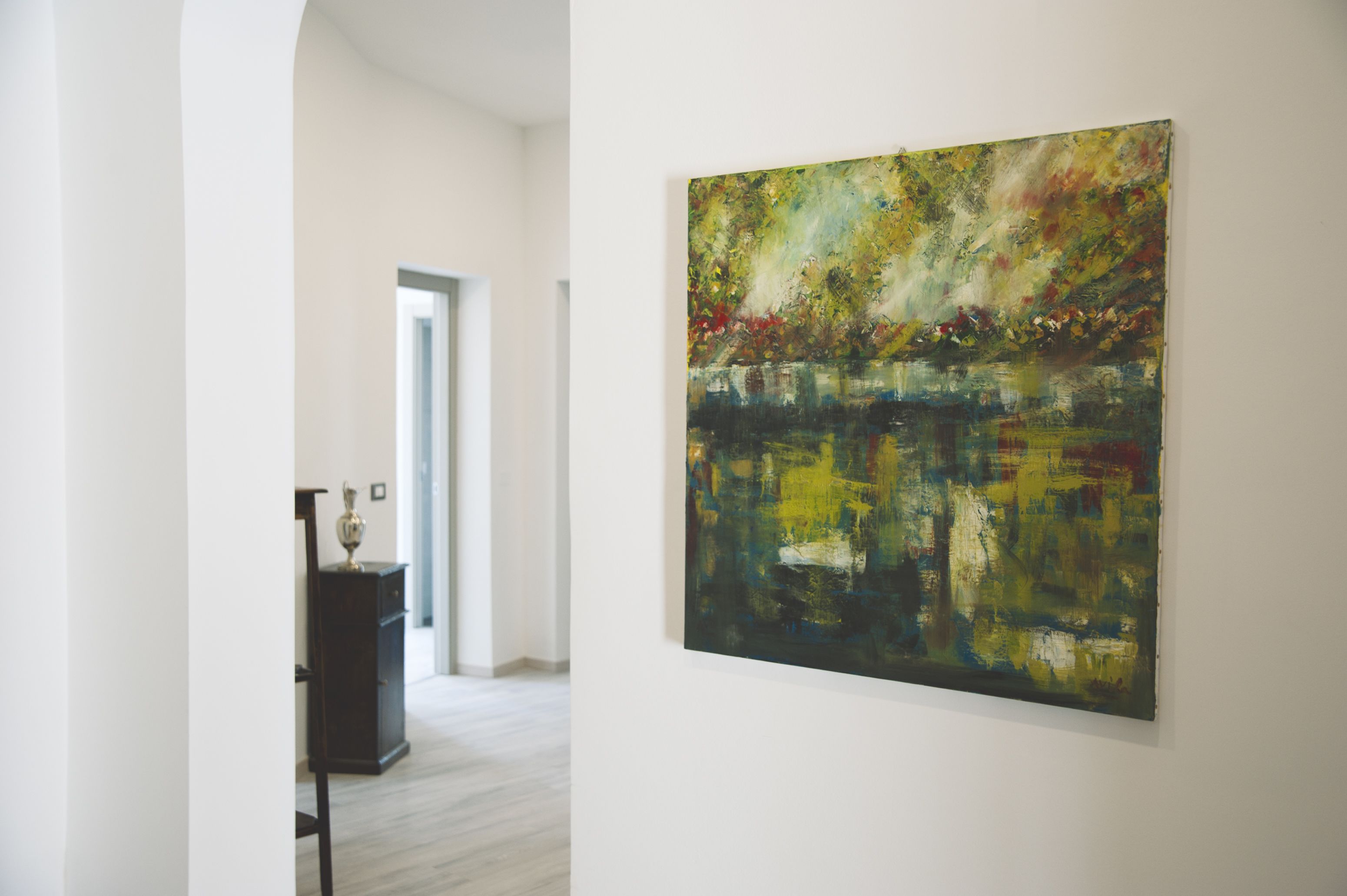 A hallway with a large abstract painting on the wall, a dark cabinet with a vase, and a door leading to another room.