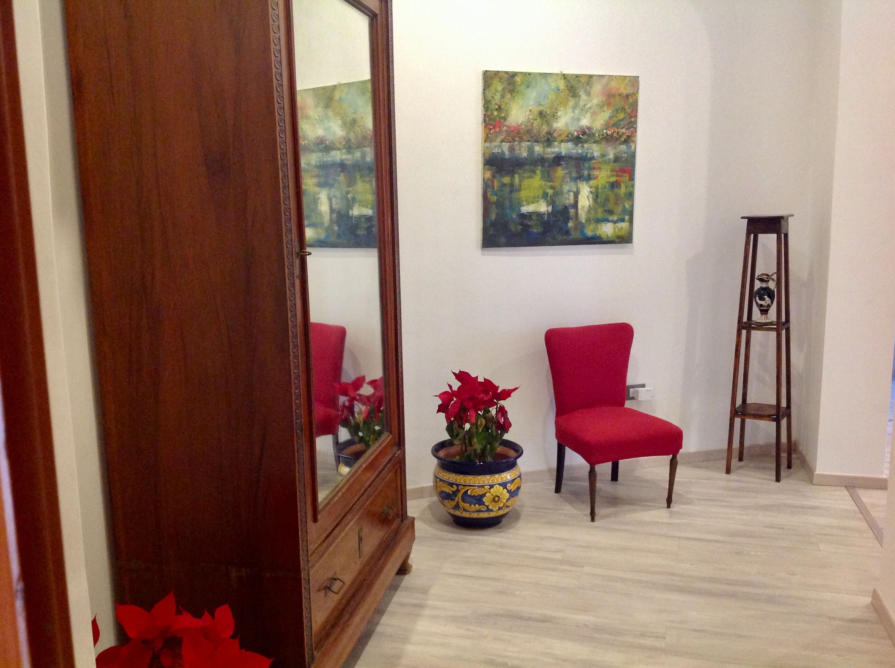 A room with a wooden floor, a red chair, a tall wooden stand, a large mirror, and a painting on the wall.