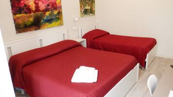 A room with two single beds, each with a red bedspread and white headboards. There is a white chair and a small table with a red cloth. Two paintings are hung on the wall.