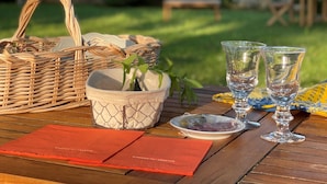 A wooden table set outdoors with a wicker basket, a small white pot with a plant, two clear glasses, and a red menu. - Domaine de Labarthe (Espere)