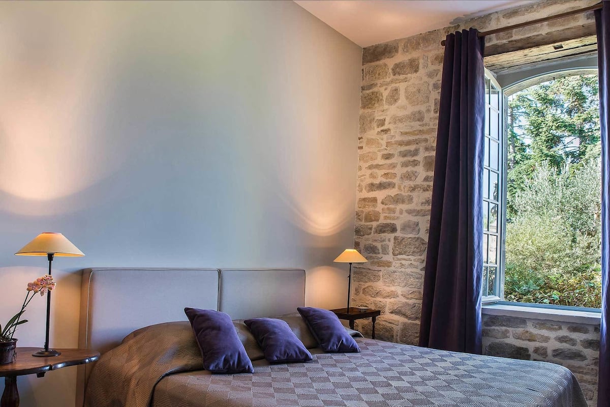 Une chambre à coucher avec un lit, deux lampes de chevet, une fenêtre avec des rideaux et une vue sur la verdure à l’extérieur.