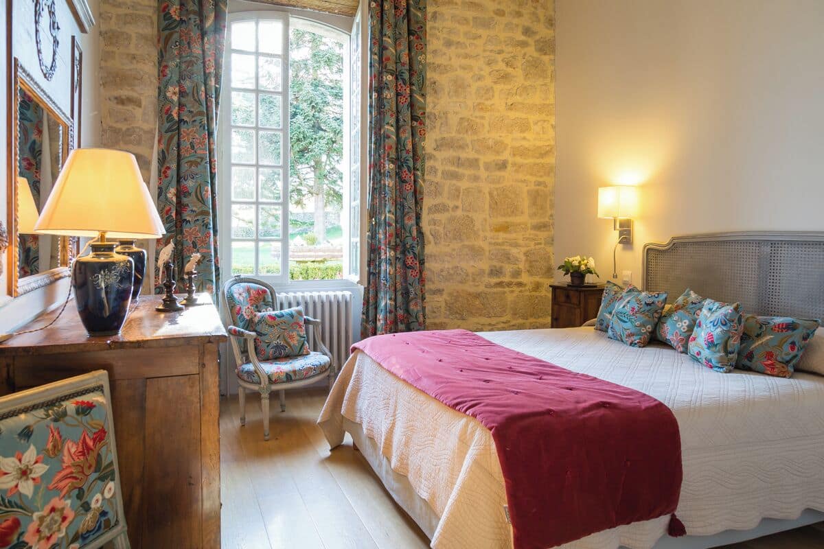 Une chambre à coucher avec un grand lit, une chaise, un bureau avec une lampe et une fenêtre avec des rideaux.