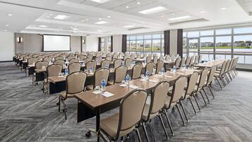 A conference room with rows of tables and chairs, large windows, and a projector screen.