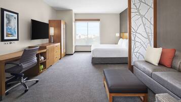 A hotel room with a bed, a desk with a chair, a sofa, a TV, and a window with a city view.