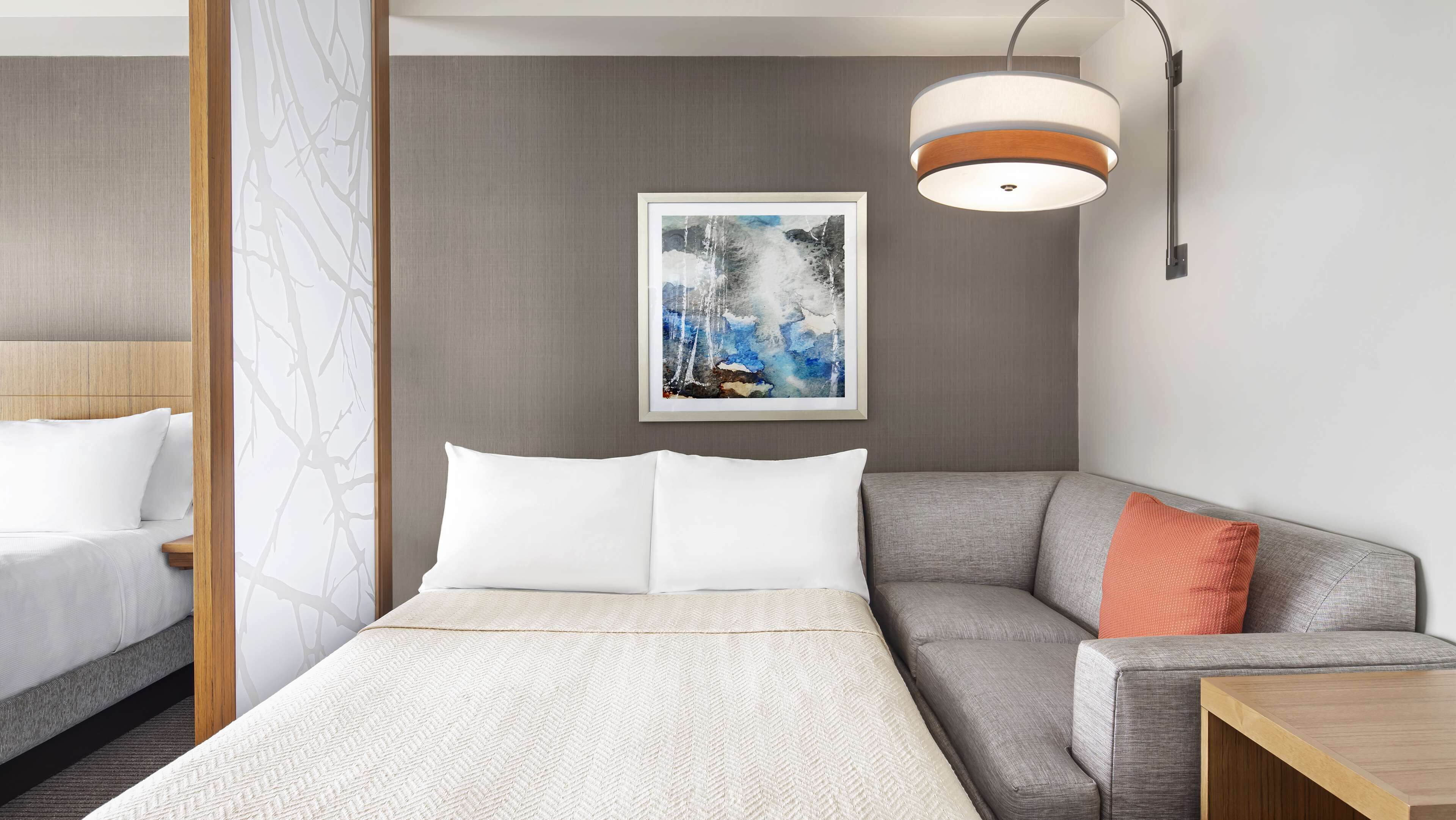 Una habitación de hotel con cama, sofá gris, almohada naranja, un cuadro enmarco y una lámpara moderna.