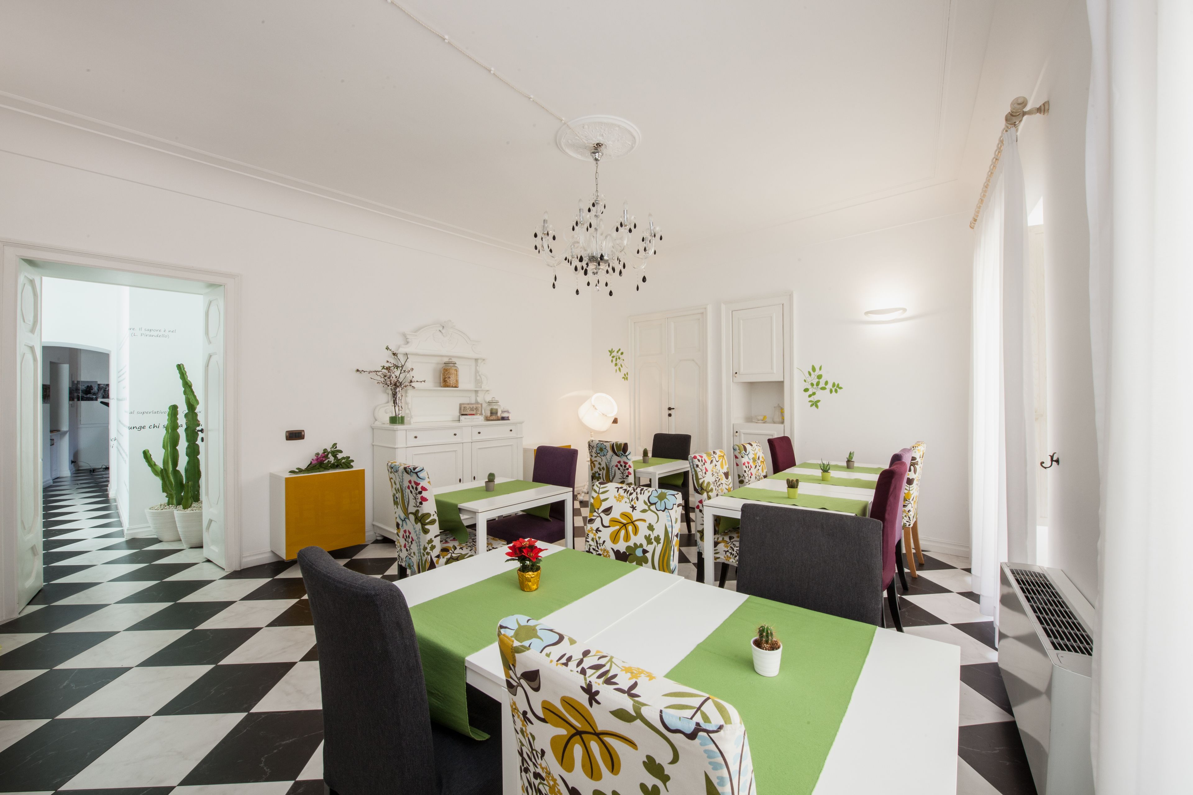 A dining area with a checkered floor, white walls, and a chandelier. There are multiple tables with chairs, some with green tablecloths.