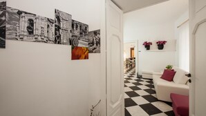 Un couloir doté d’un sol à carreaux, un mur blanc orné de photographies en noir et blanc, un canapé blanc avec des coussins rouges et une porte donnant sur une autre pièce.