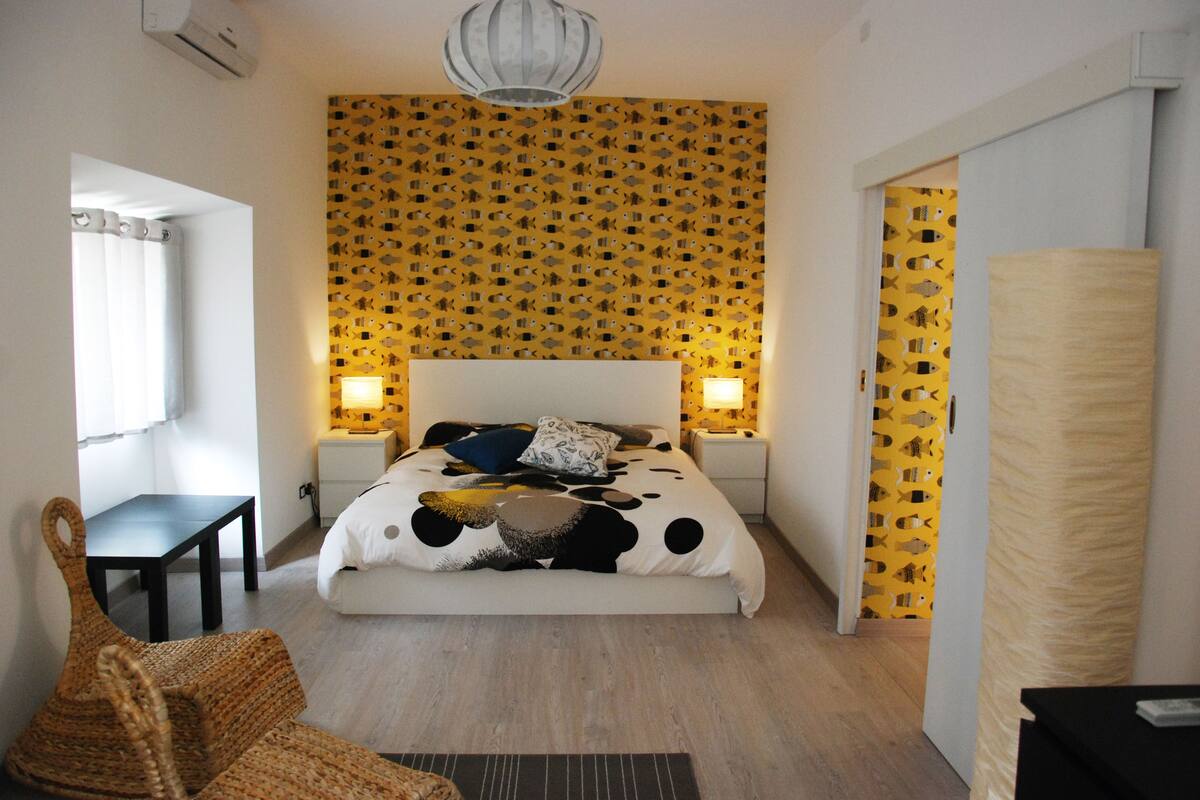 Un dormitorio con una cama, dos mesitas de noche, una silla y un papel tapiz estampado.