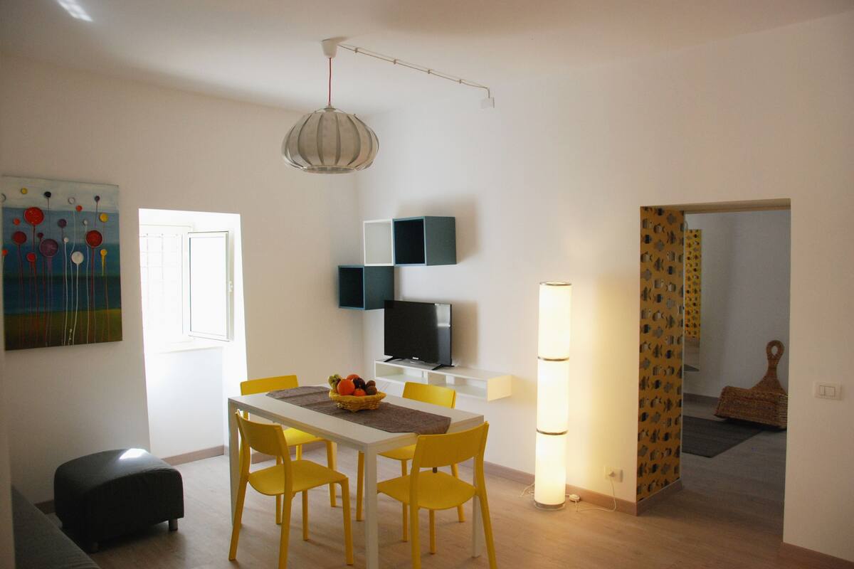 Un comedor con una mesa blanca, sillas amarillas, una cesta de frutas y un televisor sobre un mueble.