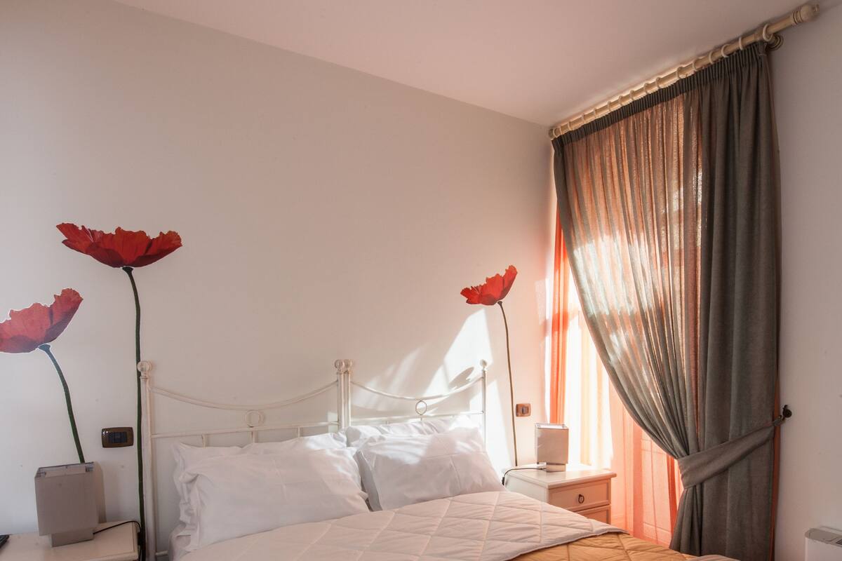 Una cama bien tendida con sábanas blancas y dos toallas sobre ella. Hay una mesita de noche con una lámpara y una pequeña mesa junto a la cama. La habitación tiene piso de madera y una ventana con cortinas.
