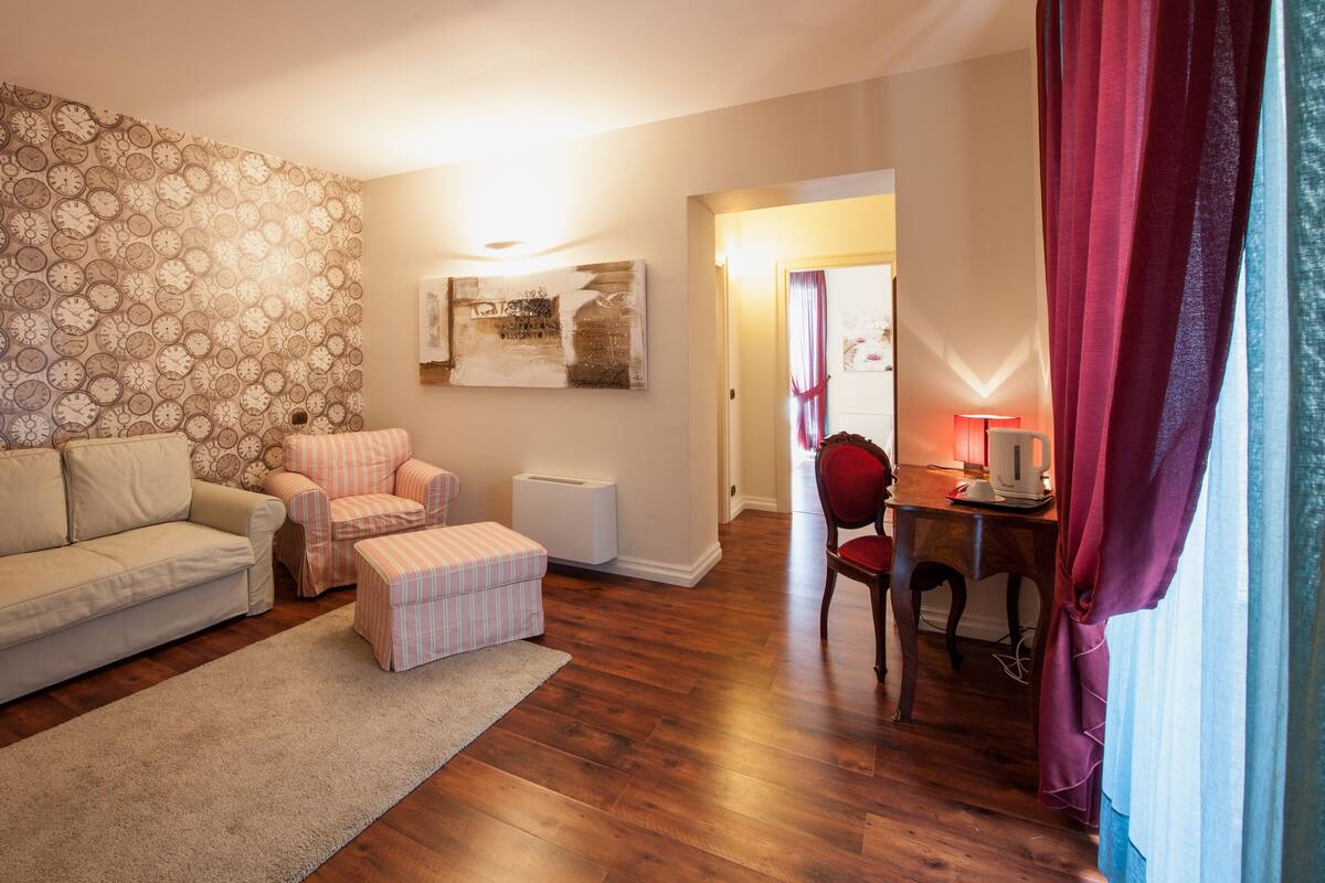 Una sala de estar con papel tapiz estampado, un sofá, un sillón, una mesita con una lámpara y un radiador.