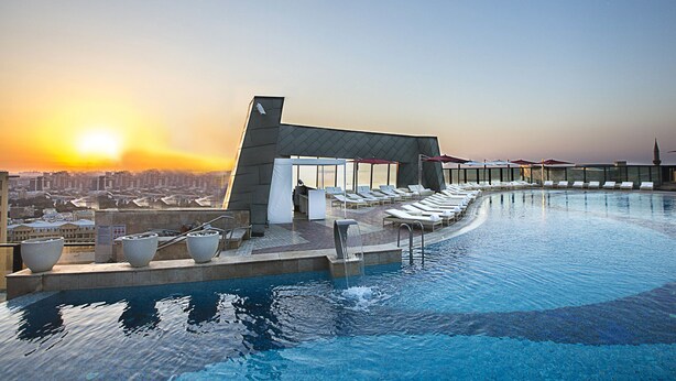 A rooftop infinity pool with a city view at sunset, lounge chairs, and a small building with a sloped roof. - Apartments by Fairmont Baku Flame Towers (Baku)