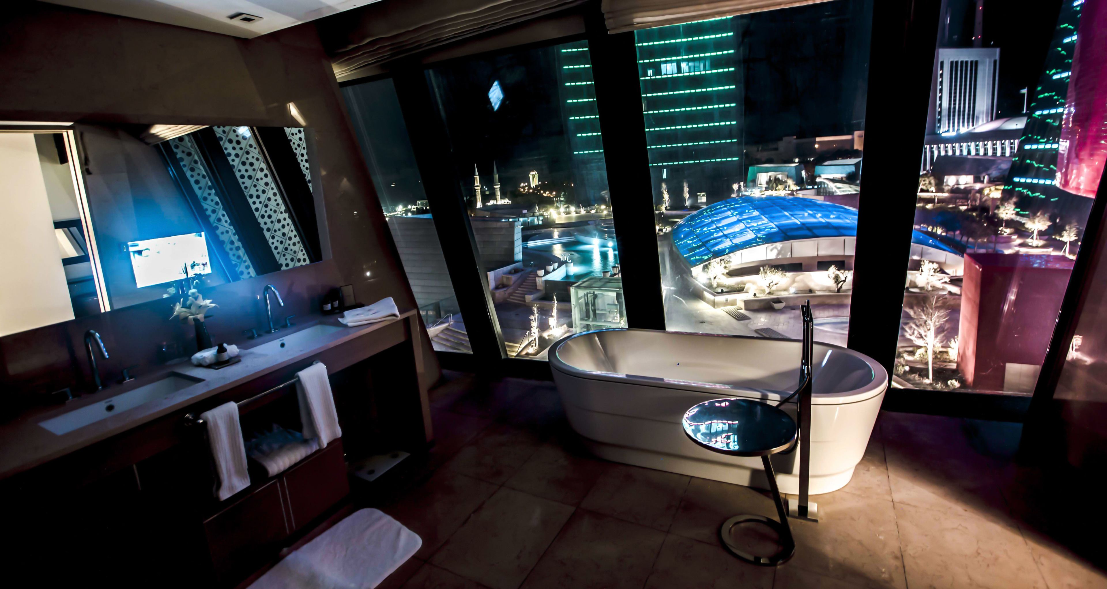 Une salle de bain moderne avec une grande baignoire, un meuble-lavabo avec un miroir et une vue sur le paysage urbain la nuit.