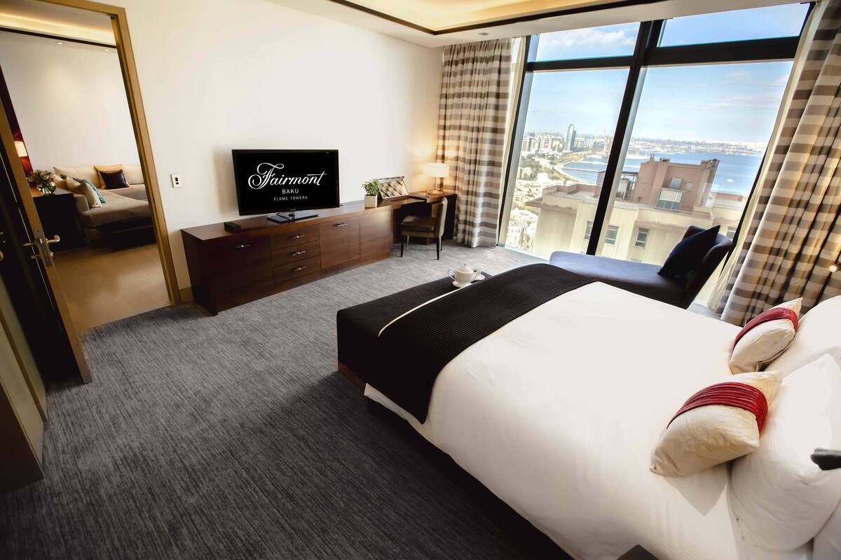 Ein Hotelzimmer mit einem großen Bett, einem Schreibtisch mit Stuhl, einem Fernseher und Blick auf die Stadt durch große Fenster.
