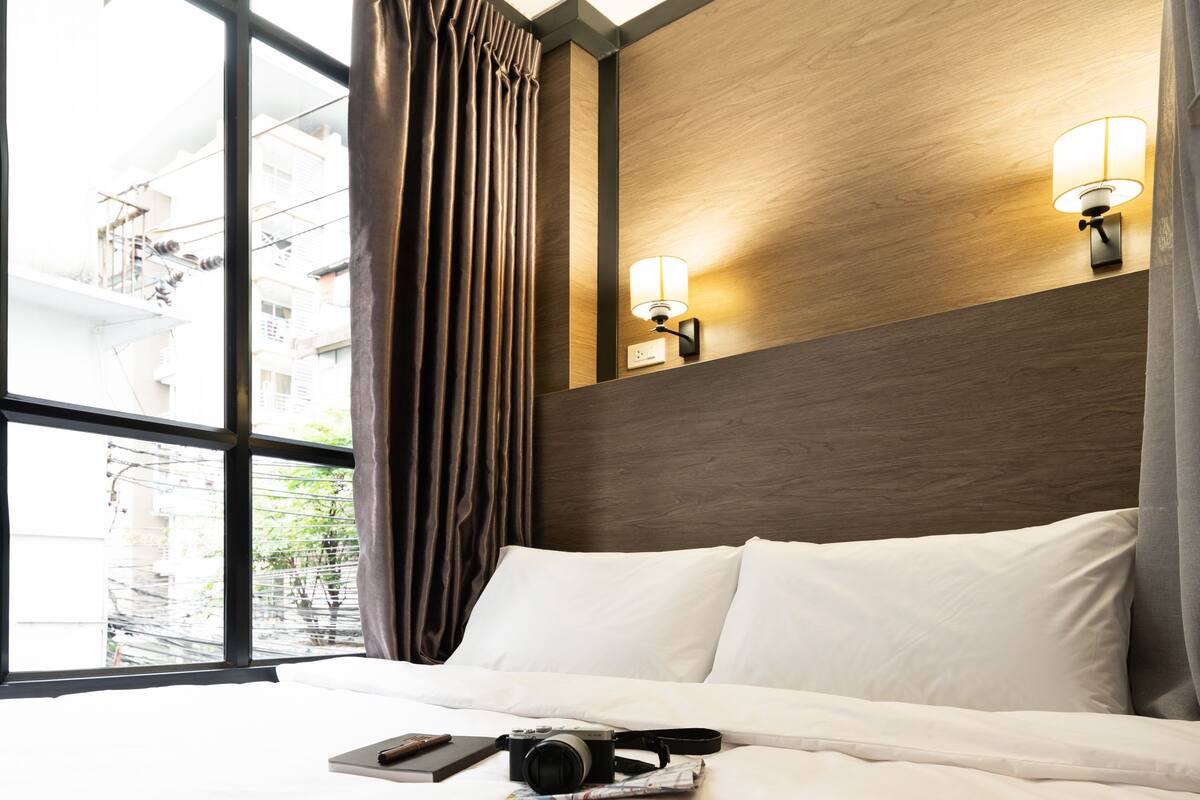 A hotel room with a large window, a bed with white linens, a camera, and a book on the bed.