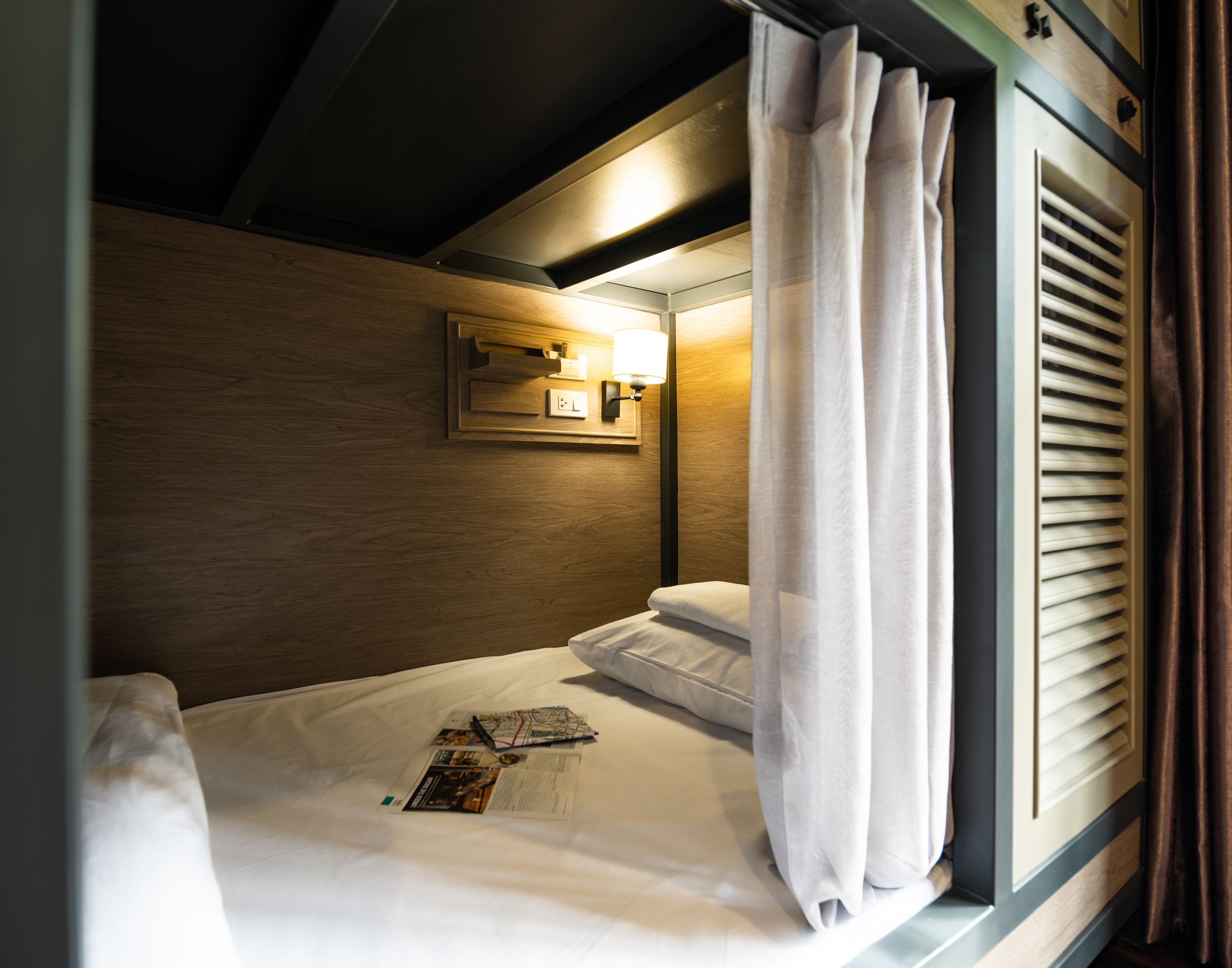 A single bed with a map and a pen, a bedside lamp, and a window with curtains.