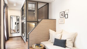A modern interior with a wooden floor, a white sofa with cushions, a wicker side table, and a staircase with glass railings.
