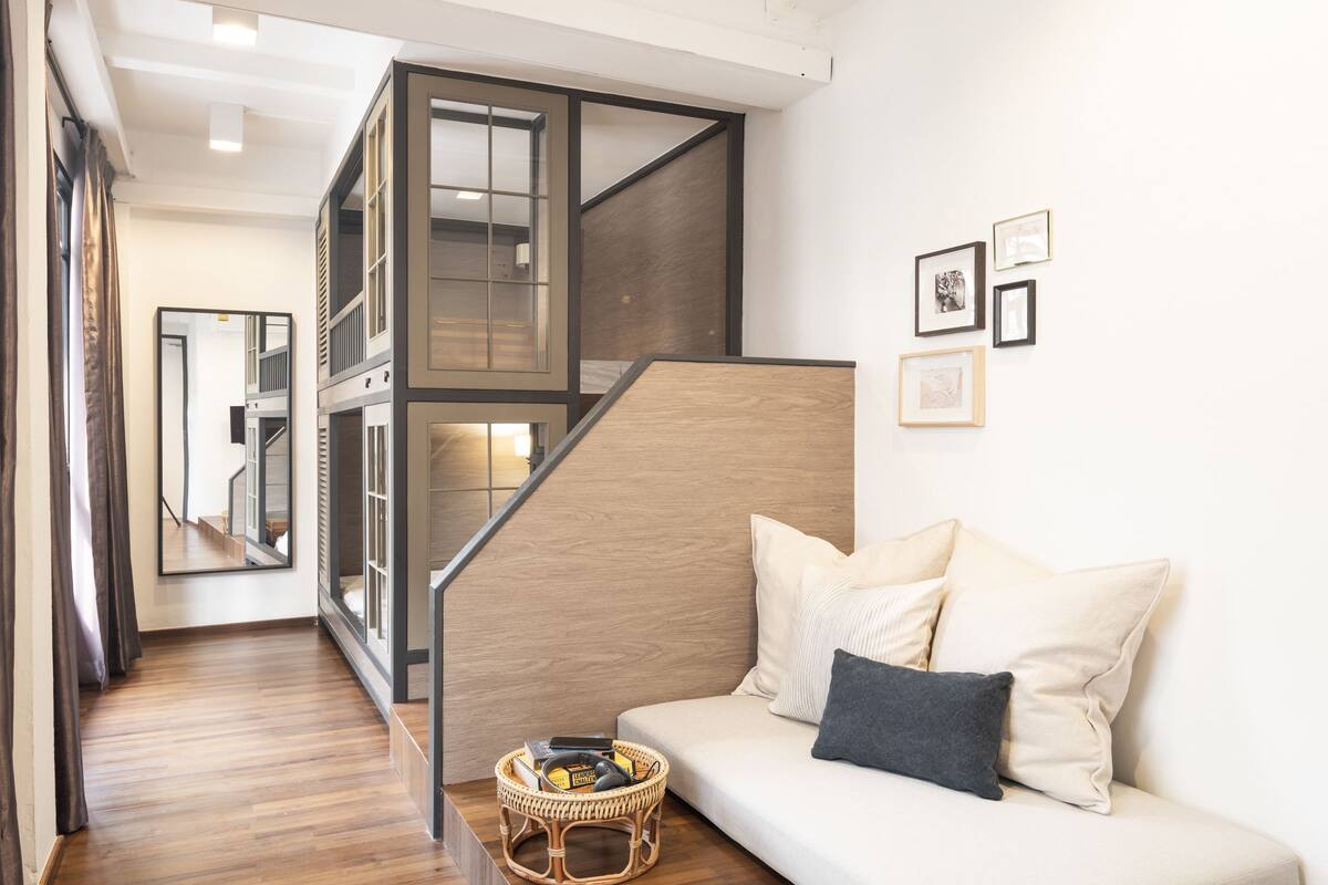 A modern interior with a wooden floor, a white sofa with cushions, a wicker side table, and a staircase with glass railings.