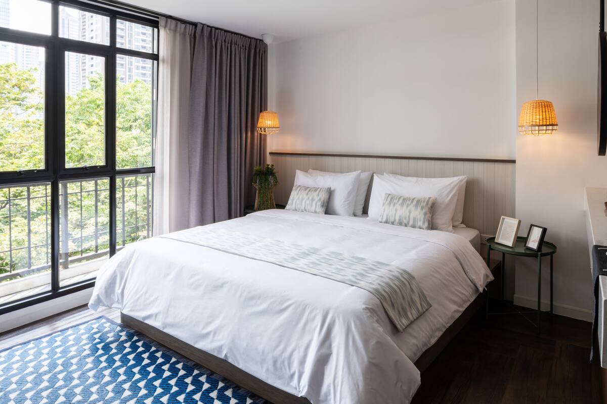 A hotel room with a large bed, a view through the window, and a small table with a lamp.