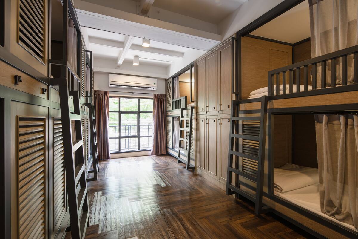 A hotel room with a bunk bed, wooden flooring, a large window with curtains, and built-in wardrobes.